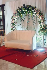 Decorative setup by Dream Tent House in Bangalore, featuring a white loveseat in front of a floral arch with white and pink flowers and greenery, placed on a red carpet.