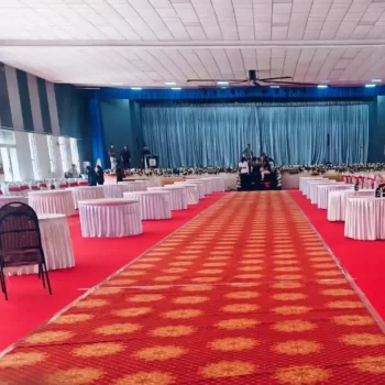 Spacious indoor event setup in Bangalore by Dream Tent House, featuring neatly arranged round tables draped in white covers, on a vibrant red carpet with a stage in the background. Serves as a prime example of elegant tent rental services in the city.