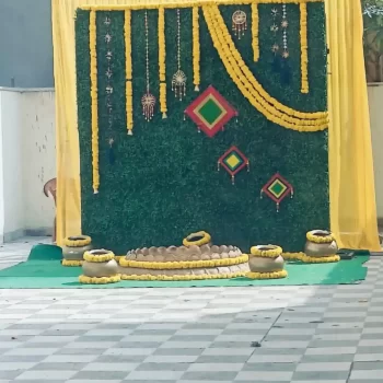 Decorative area set up by Dream Tent House in Bangalore, featuring a lush green backdrop adorned with yellow marigold garlands and traditional Indian decor elements. The floor is tiled and there are decorative pots arranged artistically. Perfect setup for events and celebrations, showcasing Dream Tent House's tent and decor rental services in Bangalore.