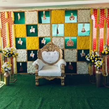 Elegant event setup by Dream Tent House, Bangalore. Features a regal chair with ornate carvings, surrounded by beautifully arranged flowers and a colourful backdrop with artistic designs. Ideal for weddings and special occasions. Tent rental services in Bangalore.