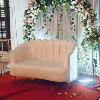 Decorative setup by Dream Tent House in Bangalore, featuring a white loveseat in front of a floral arch with white and pink flowers and greenery, placed on a red carpet.