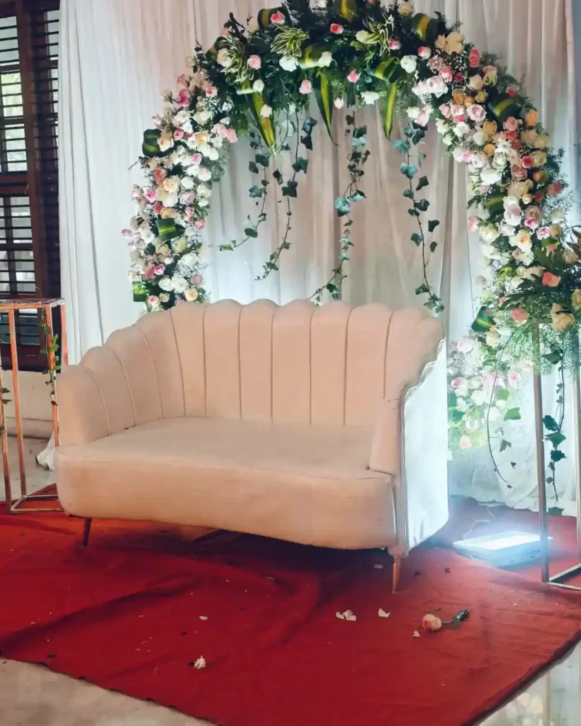 Decorative setup by Dream Tent House in Bangalore, featuring a white loveseat in front of a floral arch with white and pink flowers and greenery, placed on a red carpet.